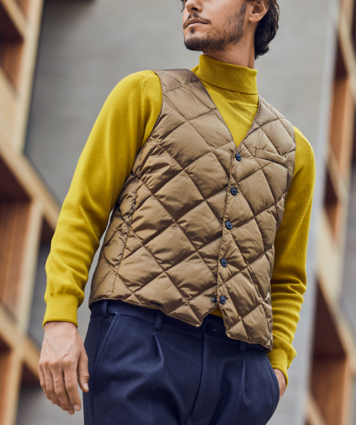 チャコールグレーのパンツ・ベージュのベスト・黄色のニットを着た外人男性