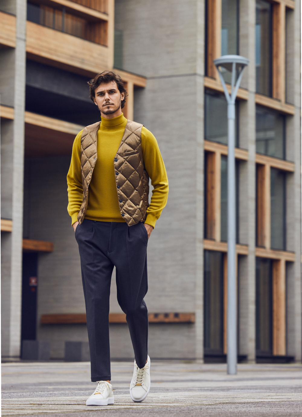 チャコールグレーのパンツ・ベージュのベスト・黄色のニットを着た外人男性