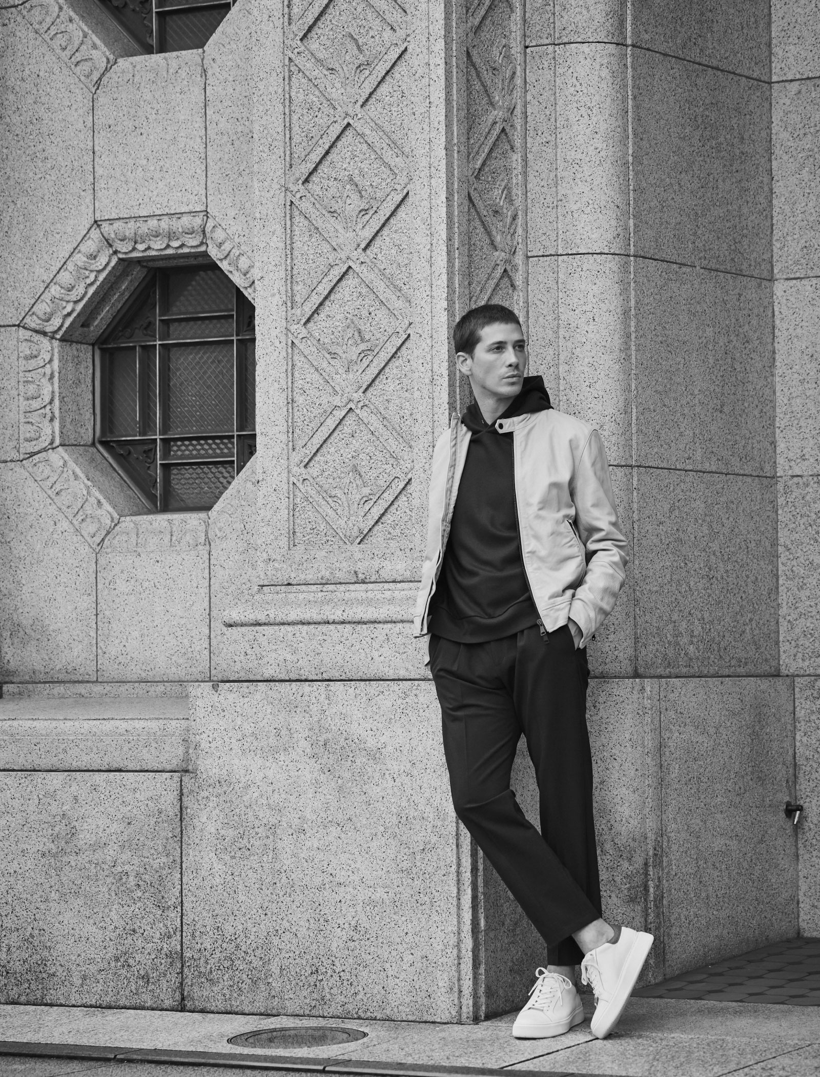 グレーの建物の壁の前に立っているアイボリーのジャケット・グリーンのパーカー・ダークグリーンのパンツ・ホワイトのスニーカーを着用したポケットに手を入れている高身長男性