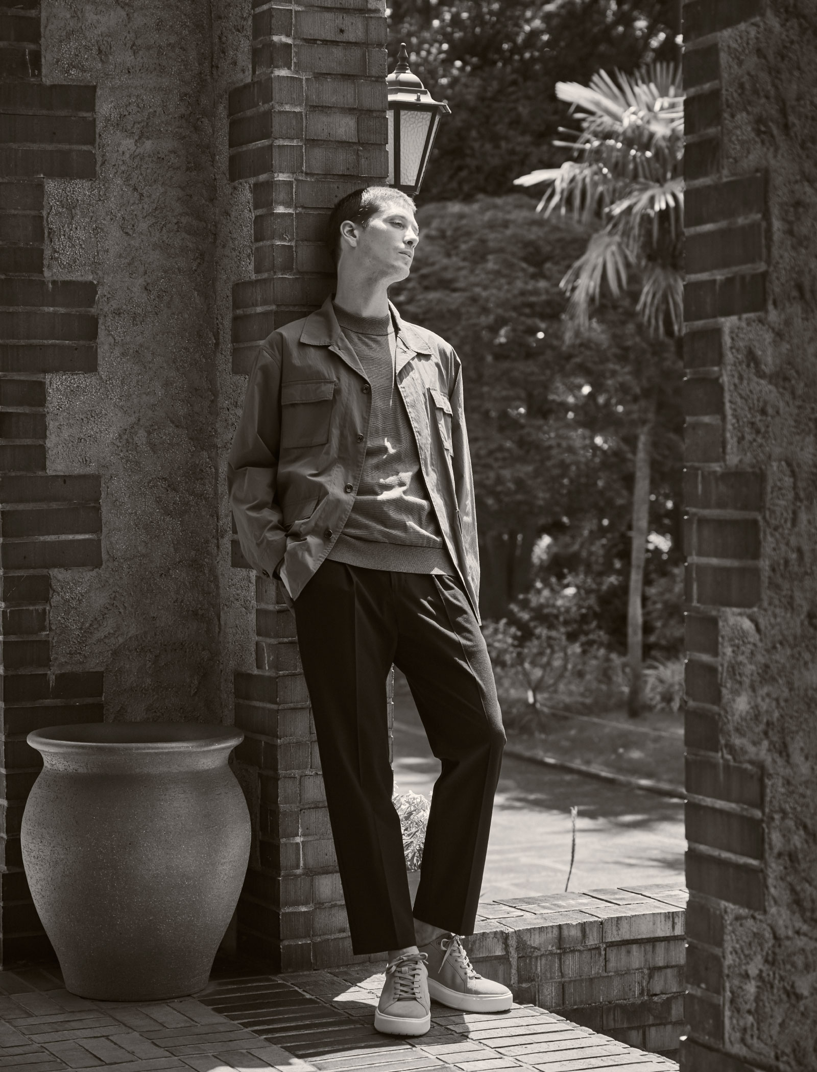 レンガでできた建物の壁に寄りかかっているグレーのアウター・チャコールグレーのシャツ・ブックのパンツ・ライトベージュのスニーカーを着用した男性