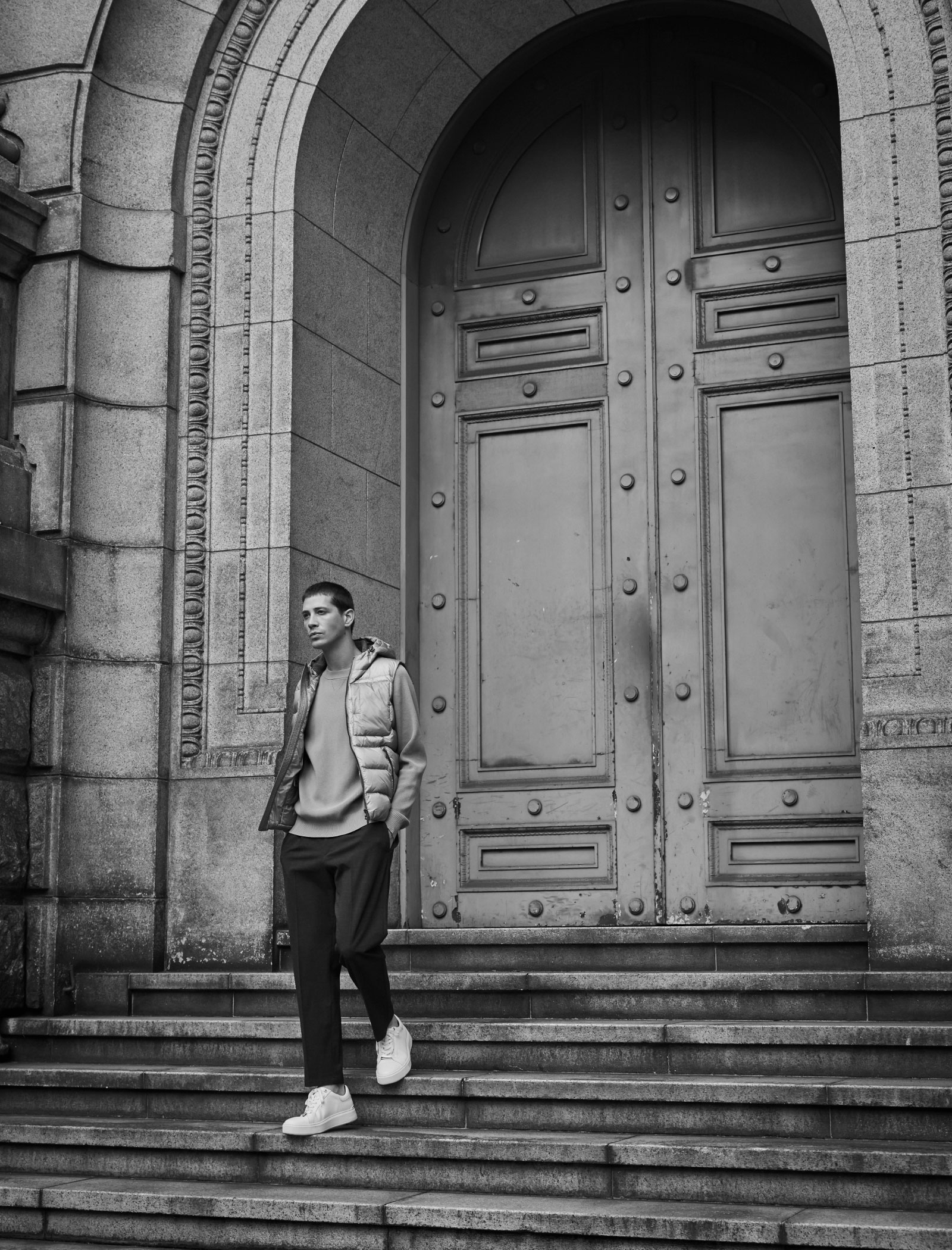 煉瓦の建物の前にいるピンクベージュのダウンベスト・ライトベージュのセーター・チャコールグレーのパンツ・オフホワイトのスニーカーを着用した高身長の男性