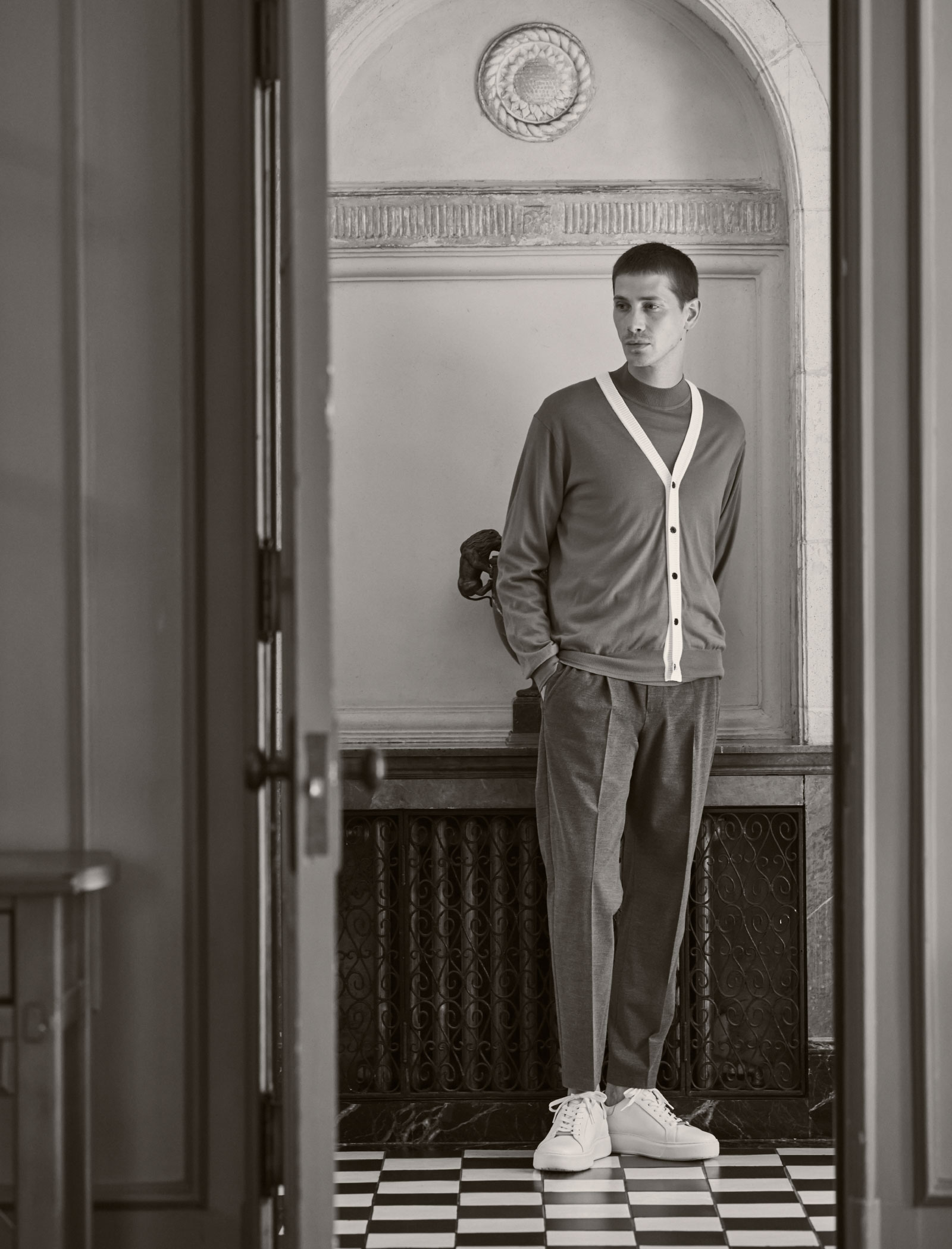 レトロな建物内の廊下にいるブルーとベージュのカーディガン・ブルーのニット・グレーのパンツ・ホワイトのスニーカーを着用した男性