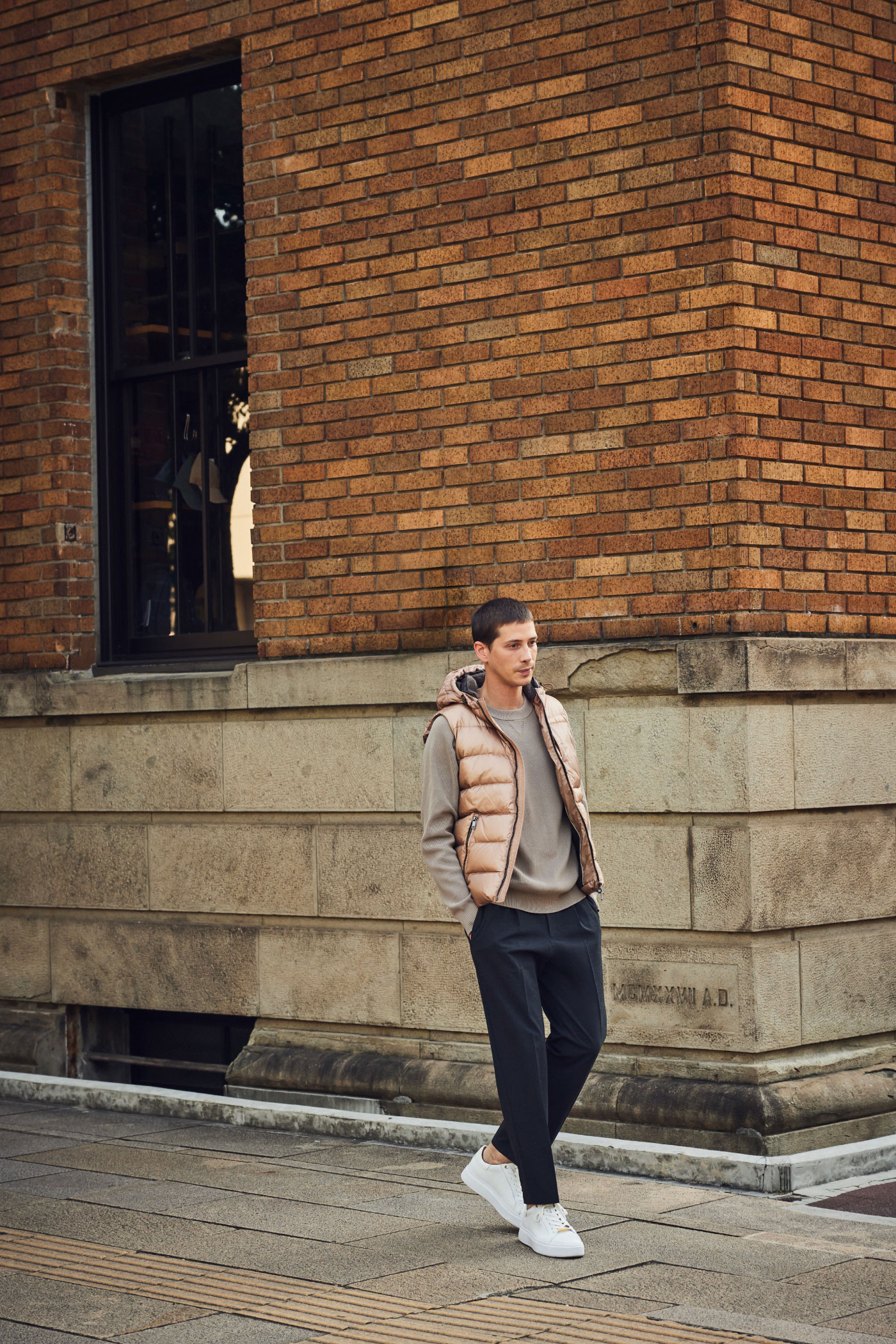 煉瓦の建物の前にいるピンクベージュのダウンベスト・ライトベージュのセーター・チャコールグレーのパンツ・オフホワイトのスニーカーを着用した高身長の男性
