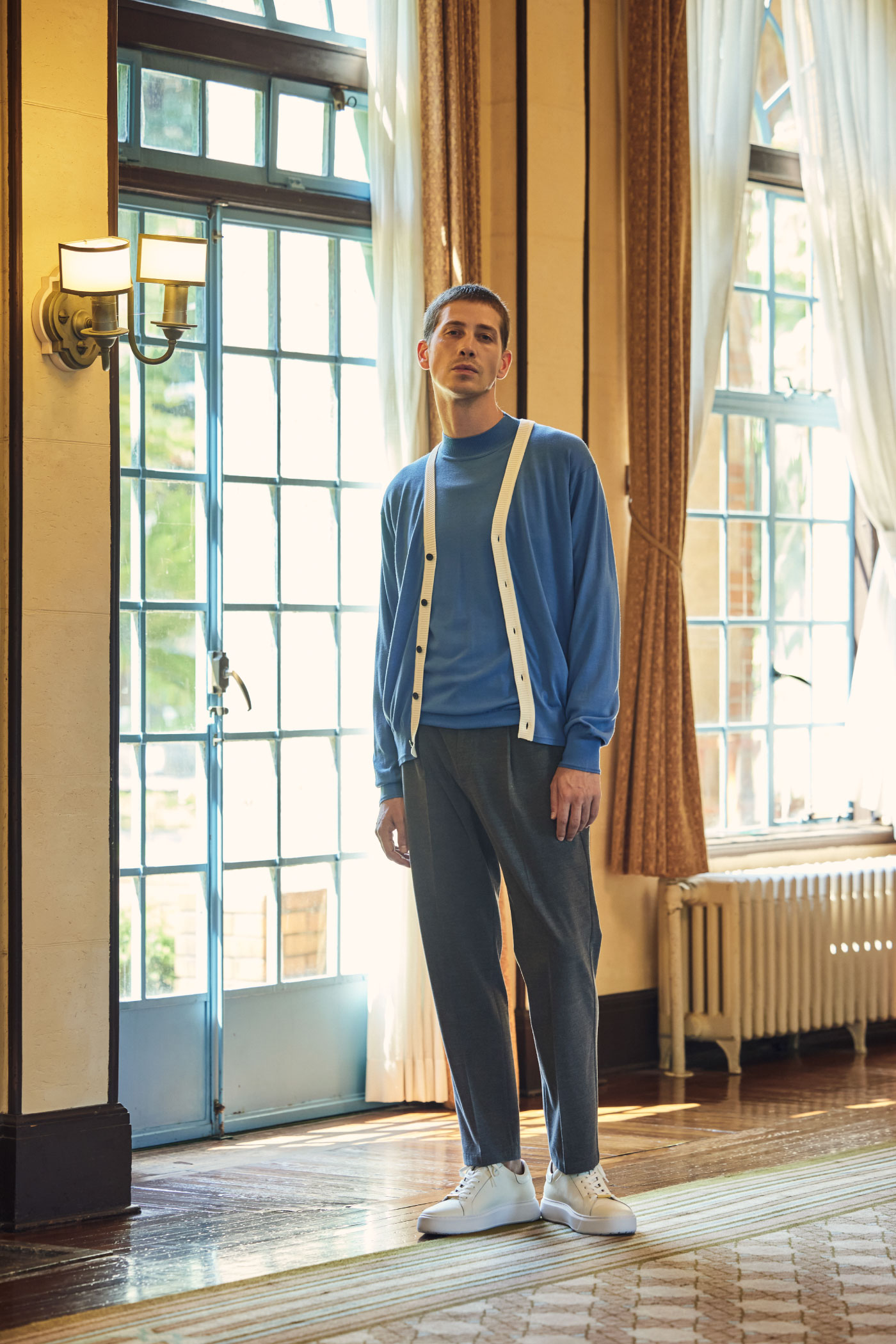 レトロな建物内の廊下にいるブルーとベージュのカーディガン・ブルーのニット・グレーのパンツ・ホワイトのスニーカーを着用した男性