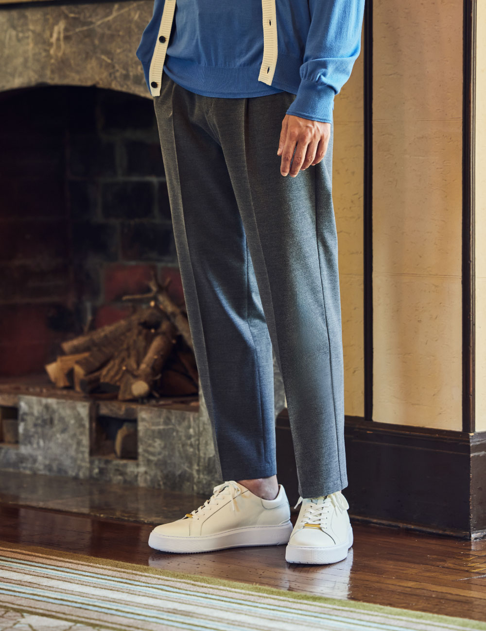 レトロな建物内の廊下にいるブルーとベージュのカーディガン・ブルーのニット・グレーのパンツ・ホワイトのスニーカーを着用した男性