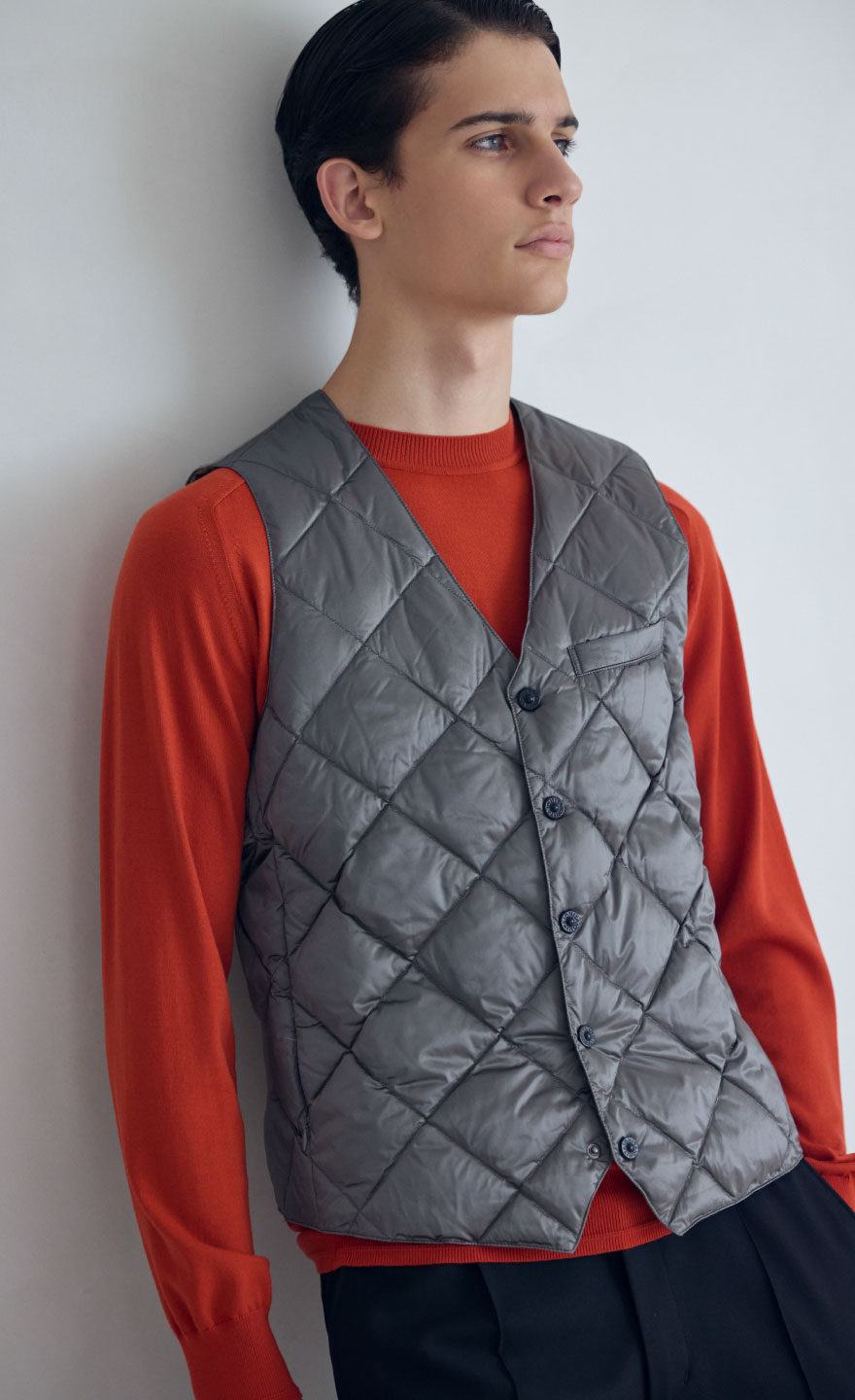 グレーのジレ・レッドのニット・ブラックのパンツ・ブラックのスニーカーを着用した、白い壁に寄りかかりながらポケットに手を入れている高身長男性