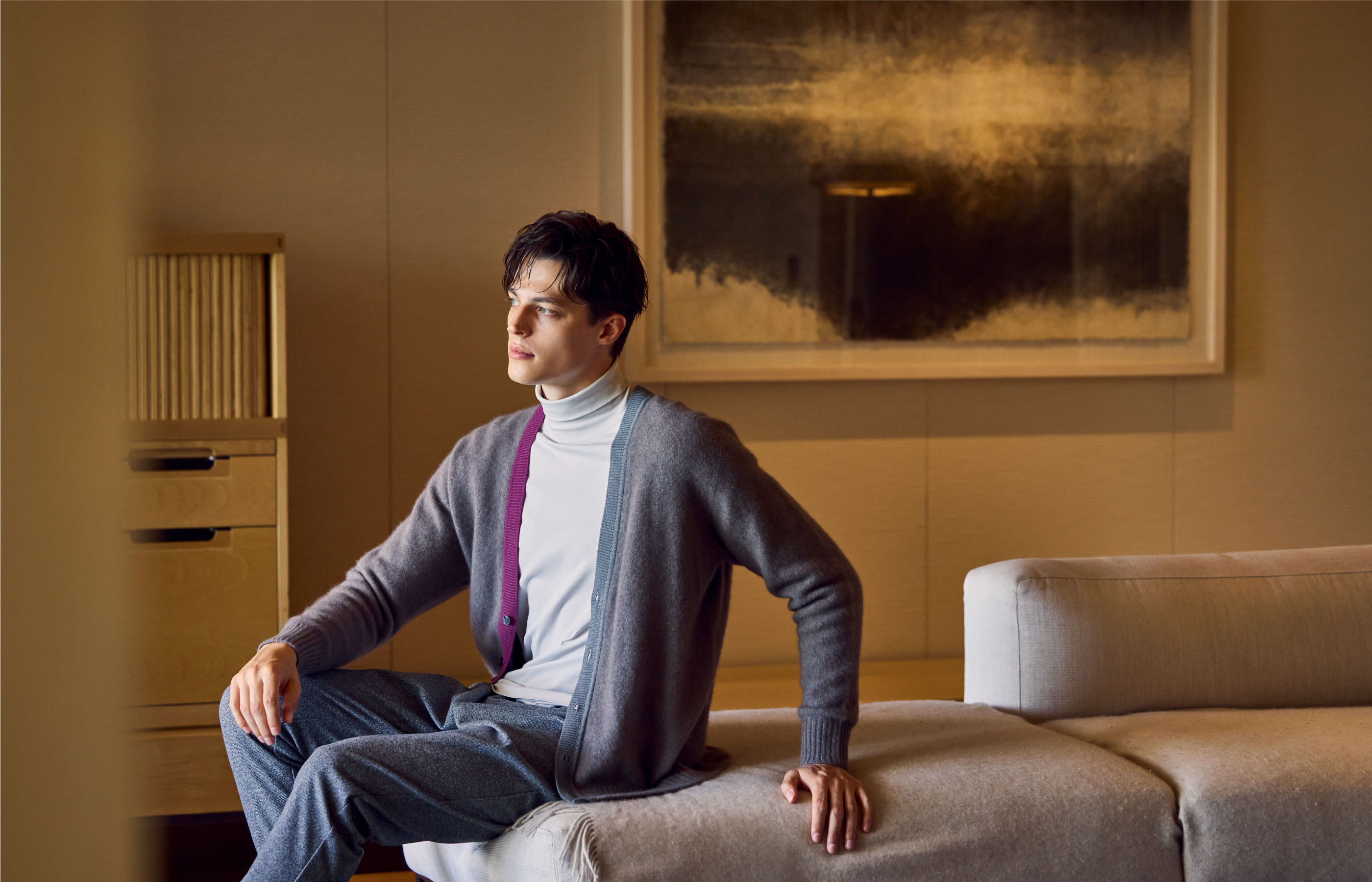 ホワイトのトップス・グレーのカーディガン・ライトネイビーのパンツを着用した、グレーのソファーに手をついて座って遠くを見ている男性