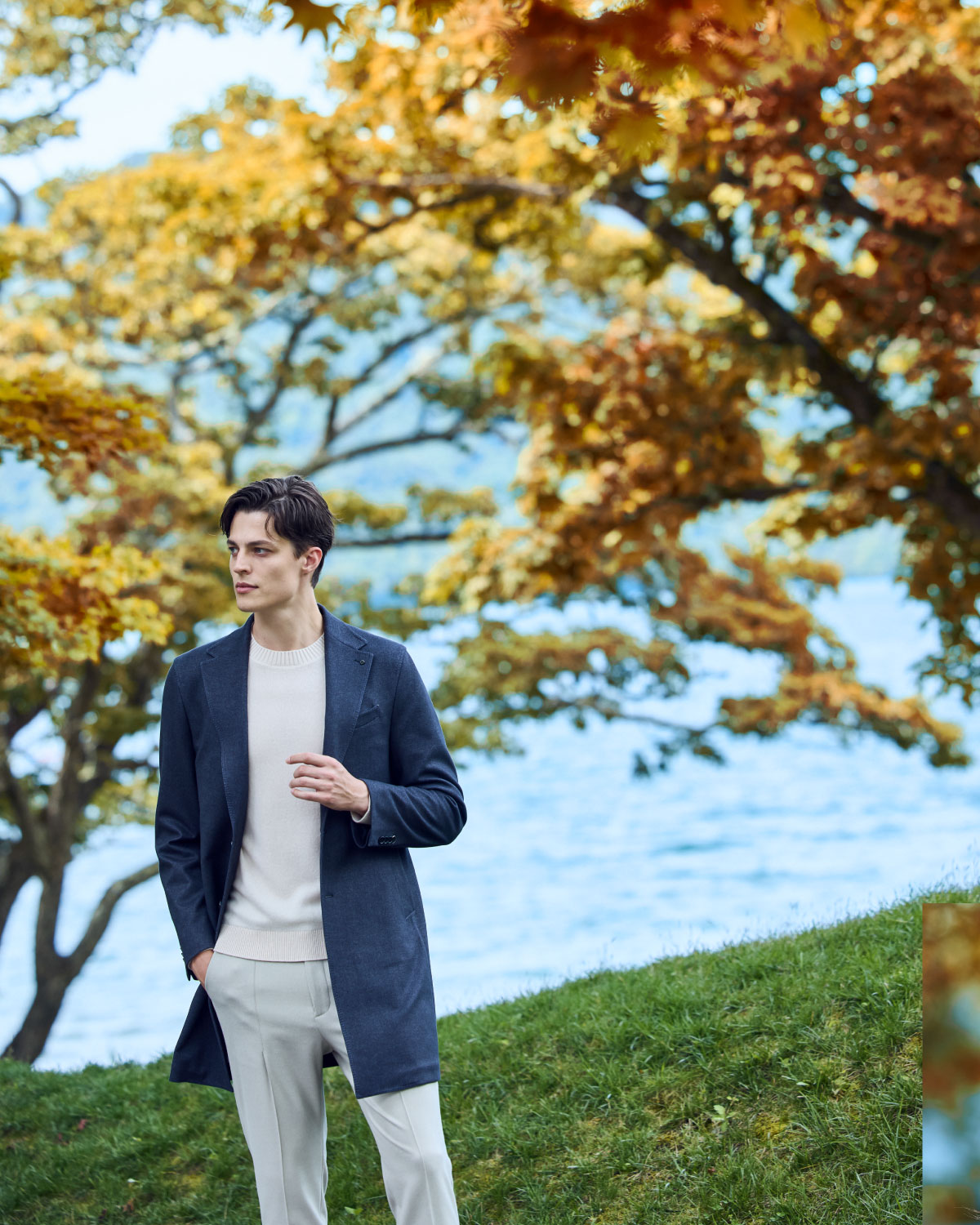ネイビーのコート・ホワイトのニット・ホワイトのパンツを着用した、芝生で手にポケットを入れて遠くを見ている男性