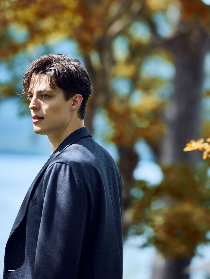 ネイビーのコート・ホワイトのニット・ホワイトのパンツを着用した、紅葉の近くで遠くを見ている男性