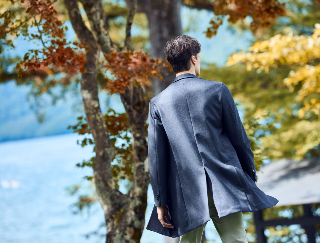 ネイビーのコート・ホワイトのニット・ホワイトのパンツを着用した、自然の中で散歩している男性の後ろ姿