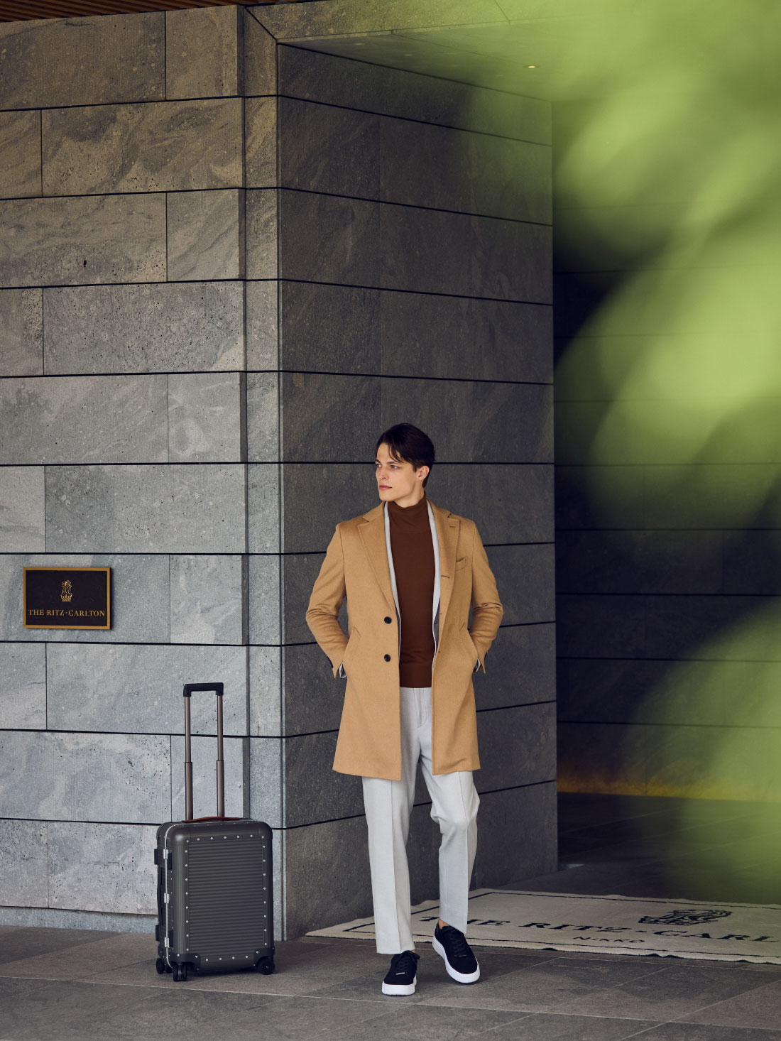 ライトベージュのコート・ホワイトのジャケット・ワインレッドのニット・ホワイトのパンツ・ブラックのスニーカーを着用した、ホテルの入り口付近で遠くを眺めている男性