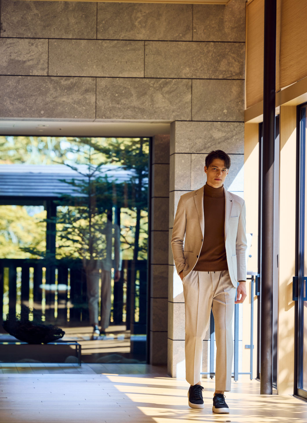ライトぐれーのジャケットセットアップ・ブラウンのニットを着用した、ポケットに手を入れて格式の高いホテルの廊下を歩いている男性