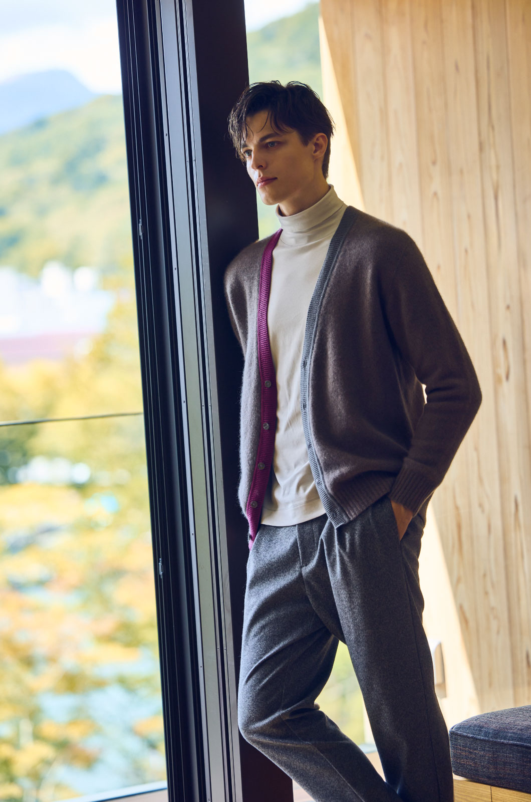 グレーのカーディガン・ライトグレーのニット・グレーのパンツを着用した、ホテルの部屋の窓のガラスによるかかっている男性