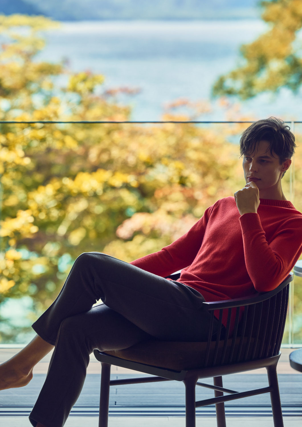 レッドのニット・ブラックのパンツを着用した、ホテルの一室のベランダでリラックスして座っている男性の様子