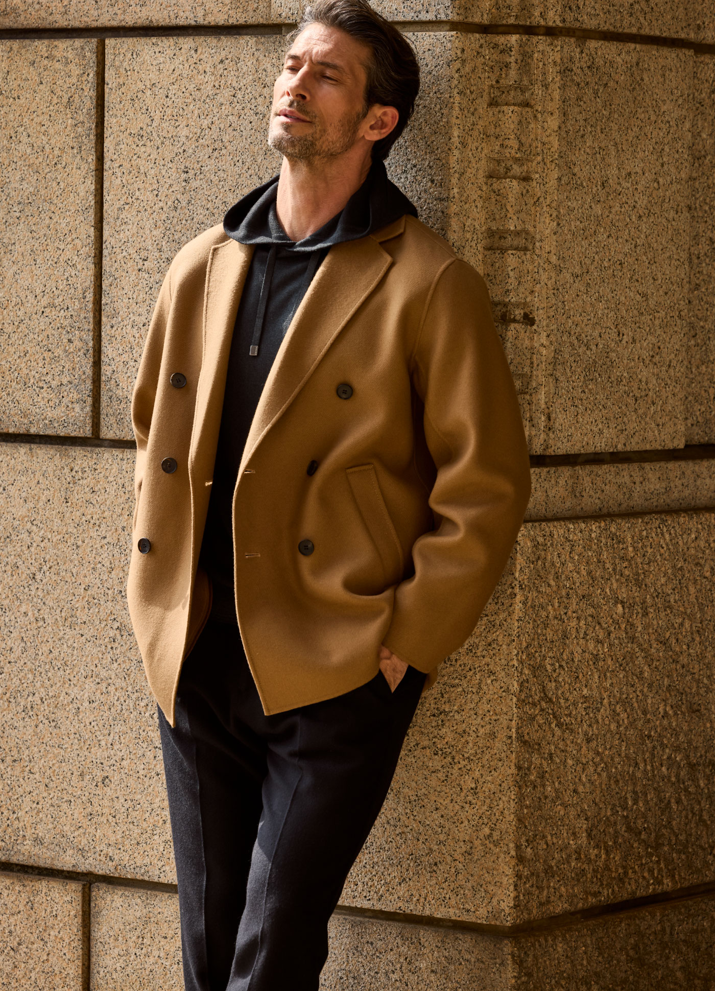 キャメルカラーのアウター・ブラックのパーカー・ブラックのパンツを着用した男性がブラウンの壁に寄りかかっている様子