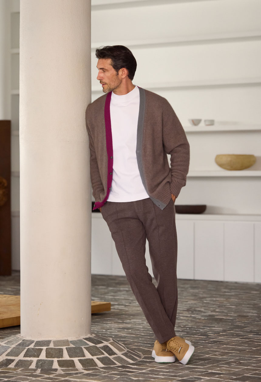 ブラウンのカーディガン・ホワイトのトップス・ブラウンのパンツ・ライトベージュのスニーカーを着用した男性がポケットに手を入れて柱に寄りかかりながら横を向いている様子