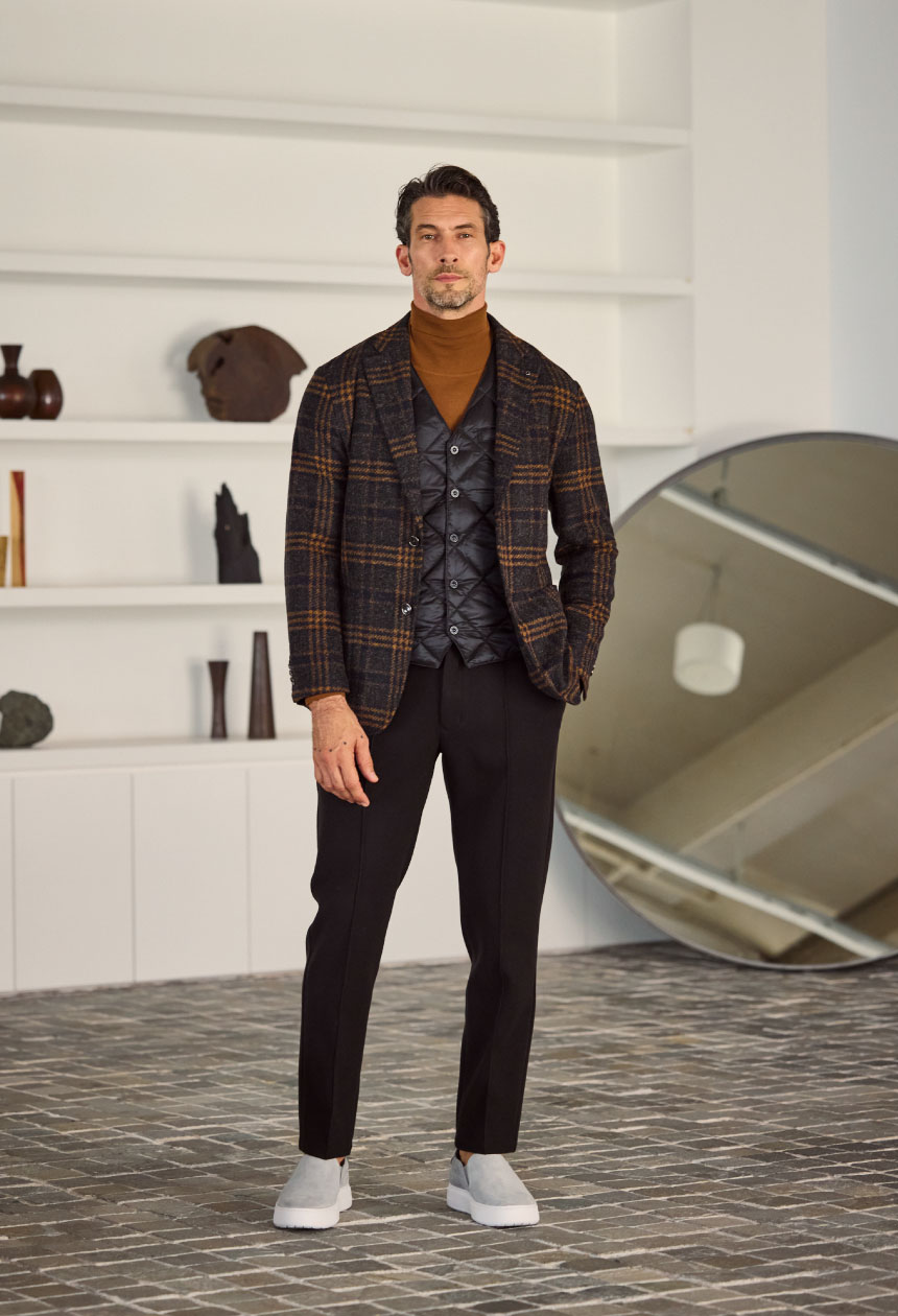 ブラウンのチェック柄のアウター・ブラックのジレ・ライトブラウンのタートルネック・ブラックのパンツ・ライトグレーのスニーカーを着用した男性がポケットに手を入れている様子