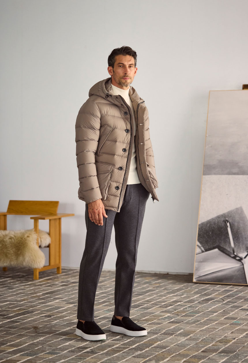 アイボリーのダウン・オフホワイトのトップス・チャコールグレーのパンツ・ブラックのスニーカーを着用した男性がポケットに手を入れて絵画の前に立っている様子