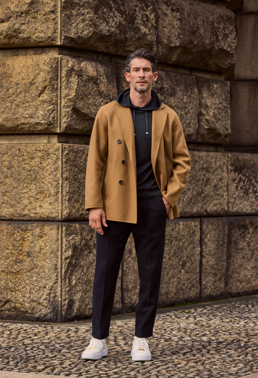 キャメルカラーのアウター・ブラックのパーカー・ブラックのパンツ・ホワイトのスニーカーを着用した男性が手をポケットに入れている様子