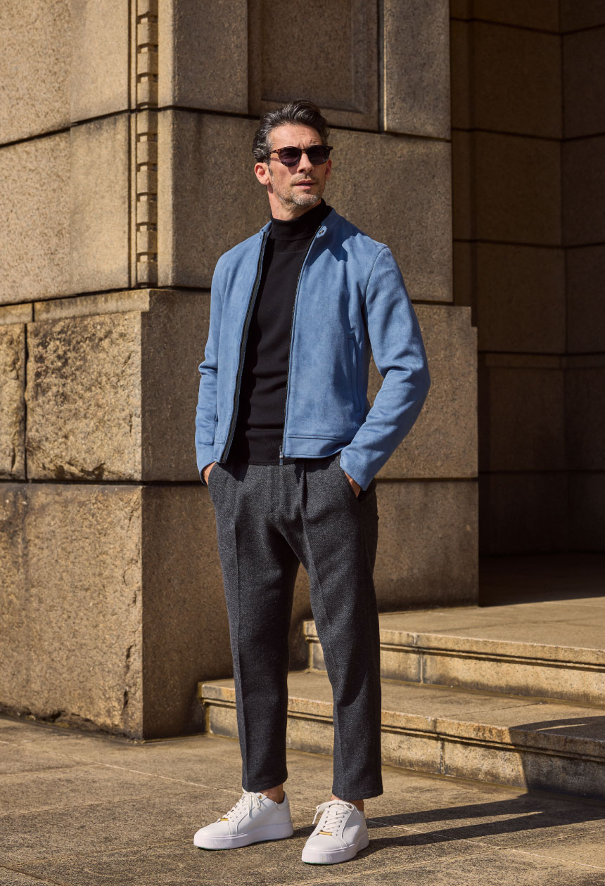 ライトブルーのライダースジャケット・ブラックのタートルネック・チャコールグレーのパンツ・ホワイトのスニーカーを着用したサングラスをかけて手にポケットを入れて遠くを見ている様子