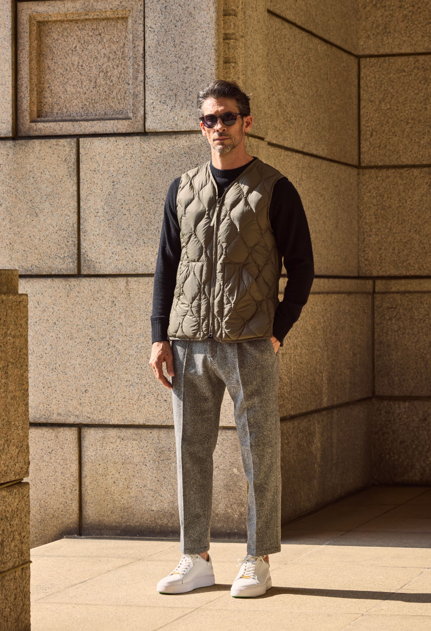 オリーブのジレ・ブラックのトップス・ライトグレーのパンツ・ホワイトのスニーカーを着用した男性がサングラスをかけてポケットに手を入れている様子