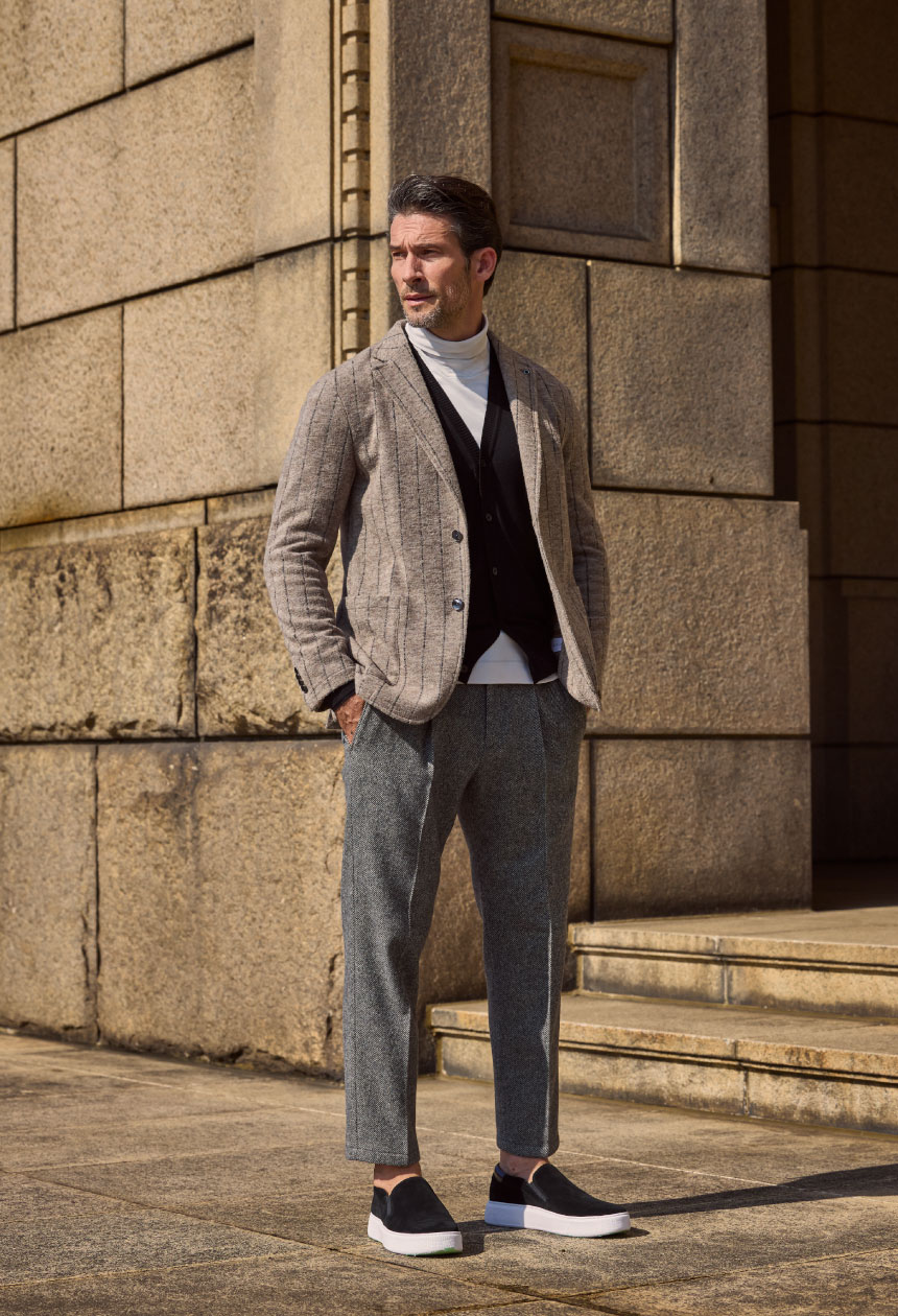 ライトベージュのジャケット・ブラックのカーディガン・ホワイトのトップス・ライトグレーのパンツ・ブラックのスニーカーを着用した男性がポケットに手を入れて横を眺めている様子