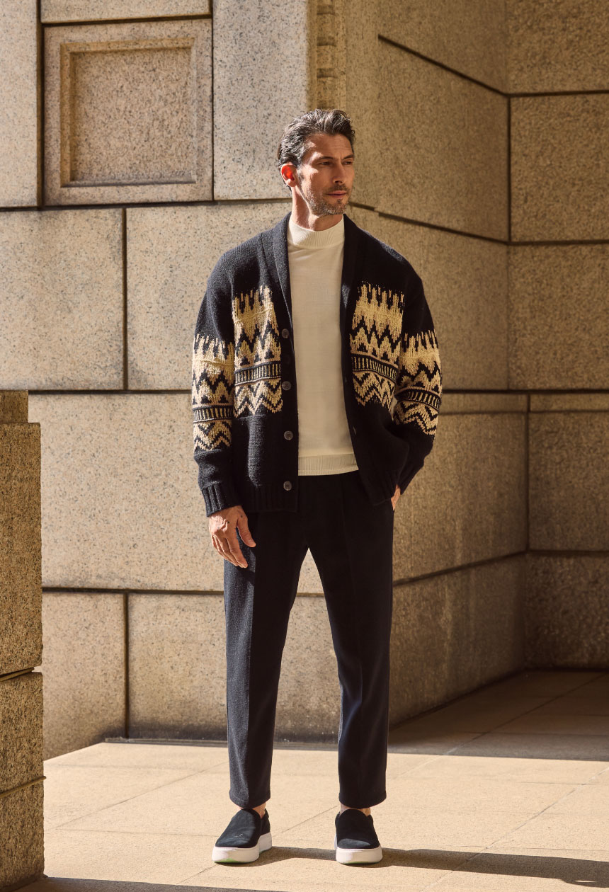 ブラックのジャケット・オフホワイトのトップス・ブラックパンツ・ブラックのスニーカーを着用した男性がポケットに手を入れている様子