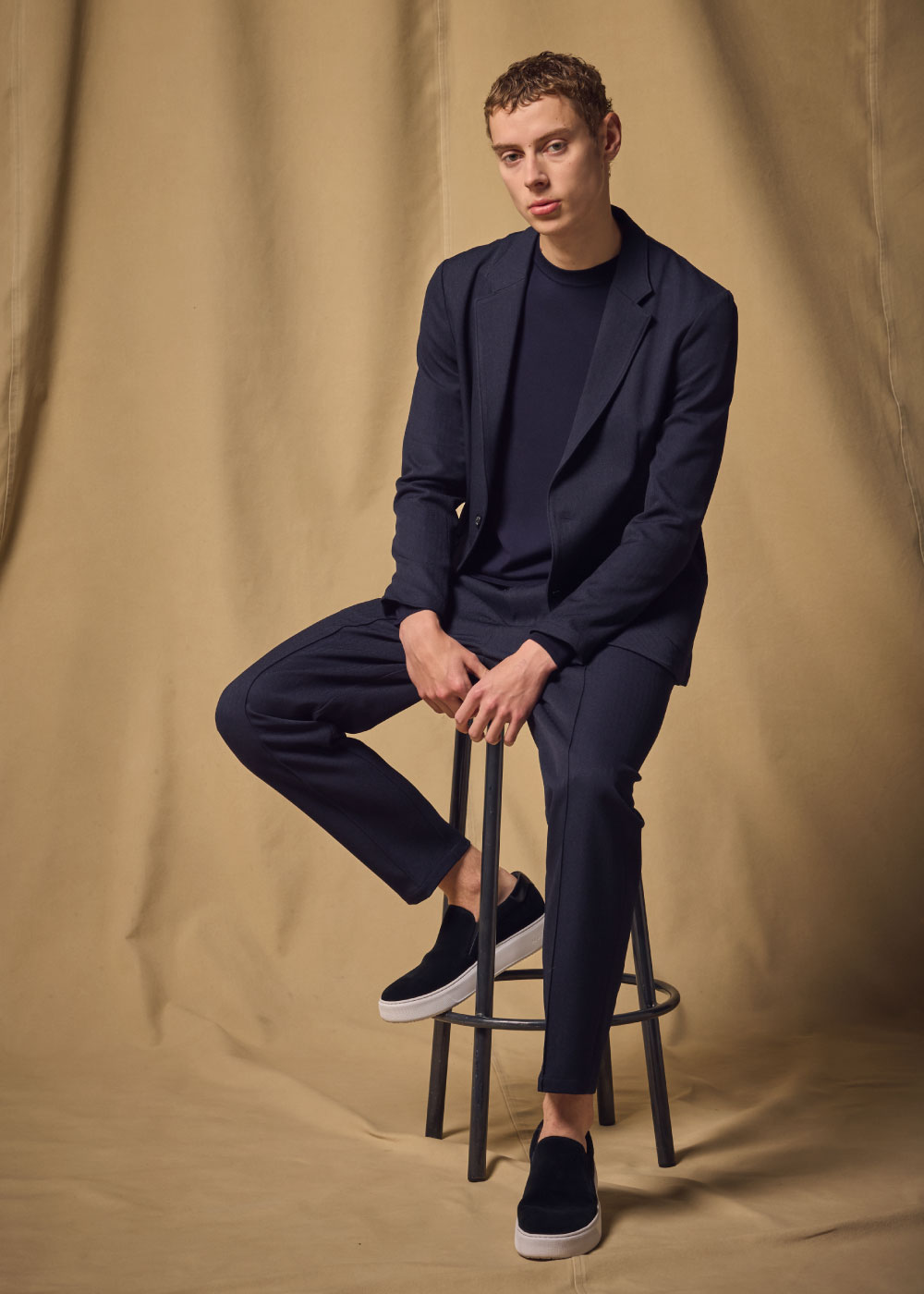 ネイビーのスーツ・ホワイトのニット・ホワイトのスニーカを着用した男性が黒い椅子に座っている様子