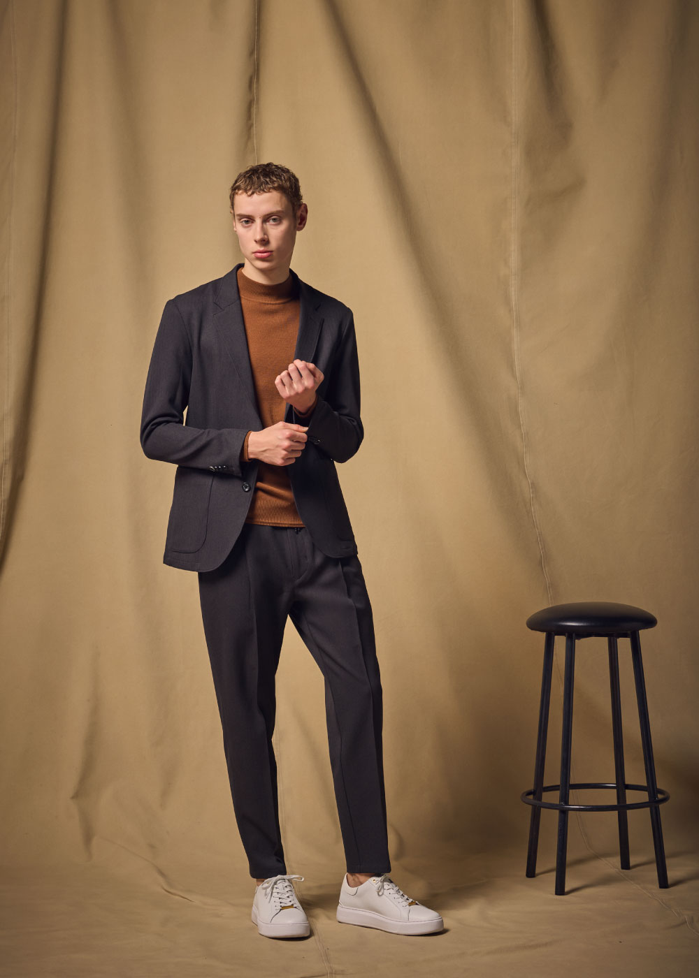 チャコールグレーのスーツ・ライトブラウンのニット・ホワイトのスニーカーを着用した男性