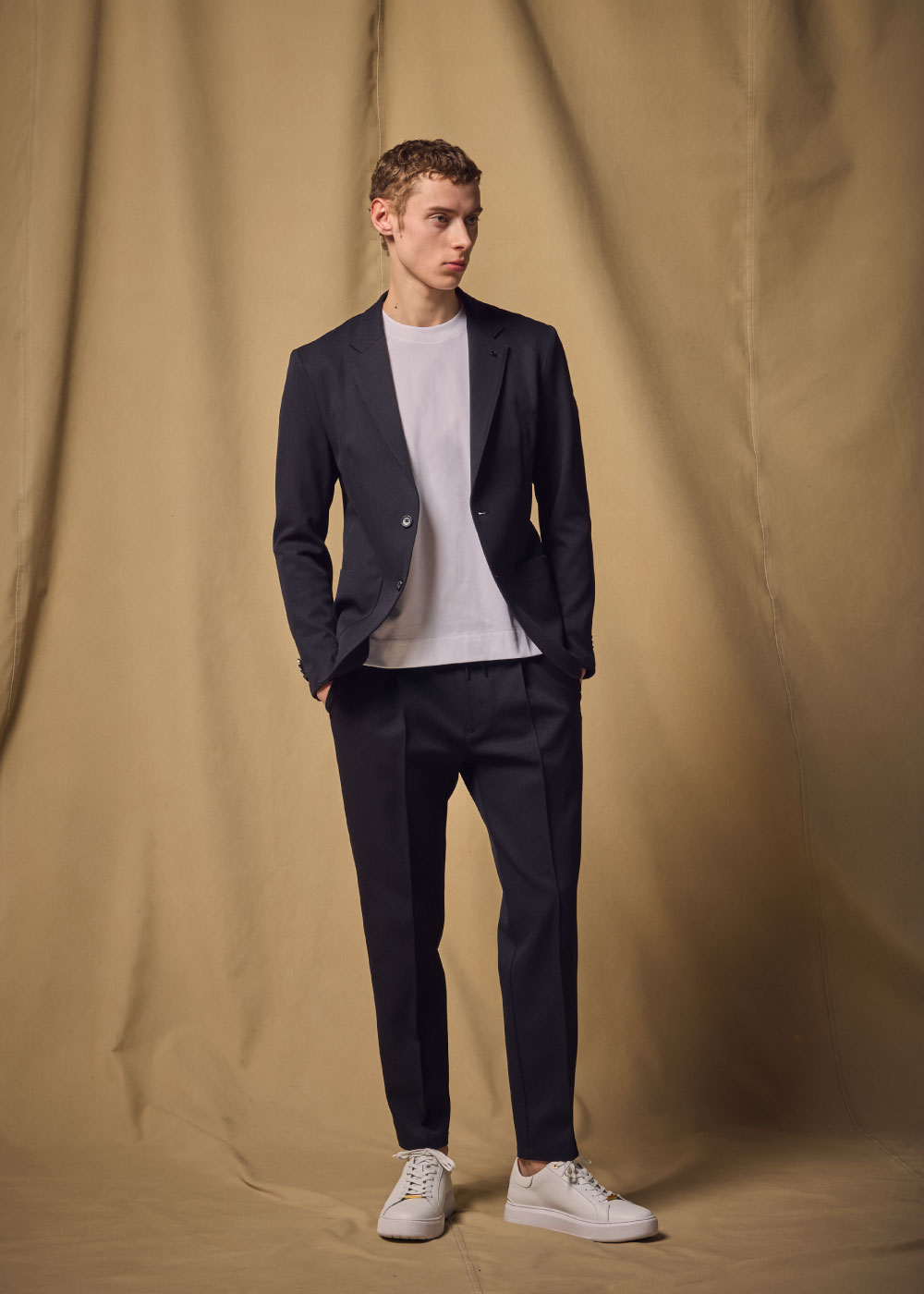 ネイビーのスーツ・ホワイトのニット・ホワイトのスニーカを着用した男性が黒い椅子に座っている様子