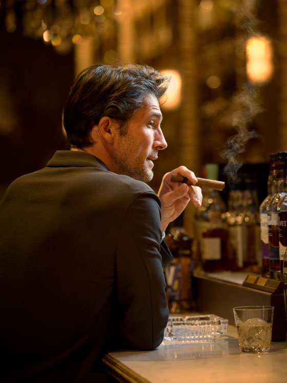 チャコールグレーのジャケット・サックスカラーのトップス・ライトベージュのパンツ・モカカラーのスリッポンを着用した男性