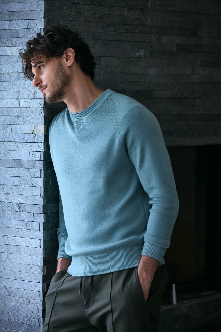 ミントのニットとカーキのパンツを着用した男性が壁にもたれかかっている画像。