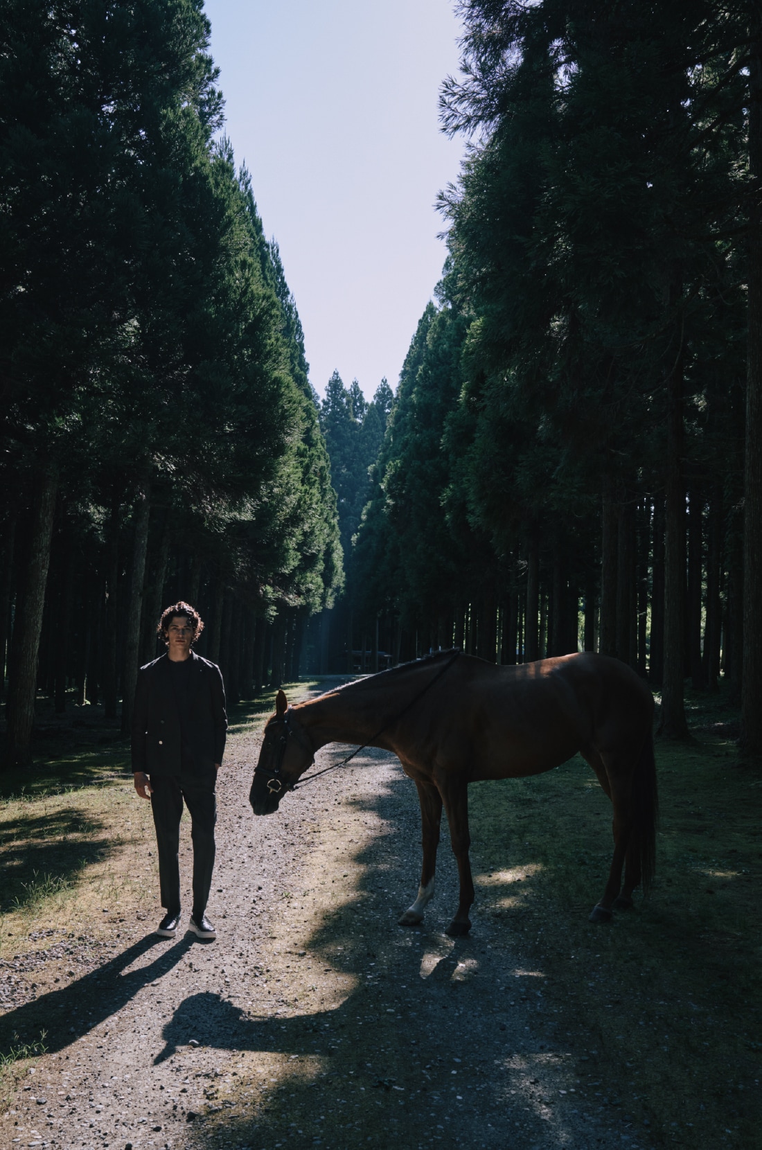 ネイビーのパックニットとブラックのスリッポンを着用して馬を連れて歩く男性のビジュアル