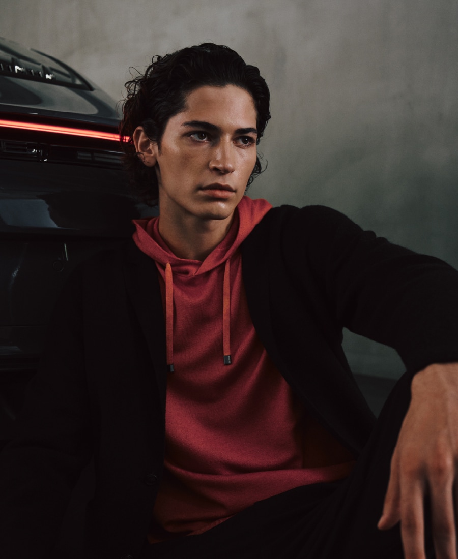 コーラルのパーカとブラックの縮絨ニットジャケット、ブラックのパンツを着用して車の前に座る男性のビジュアル