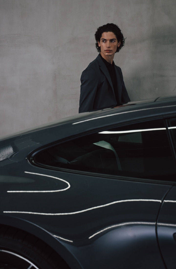 グレイのジャージーセットアップとブラックのポロシャツを着た男性が車の後ろで遠くを見つめるビジュアル