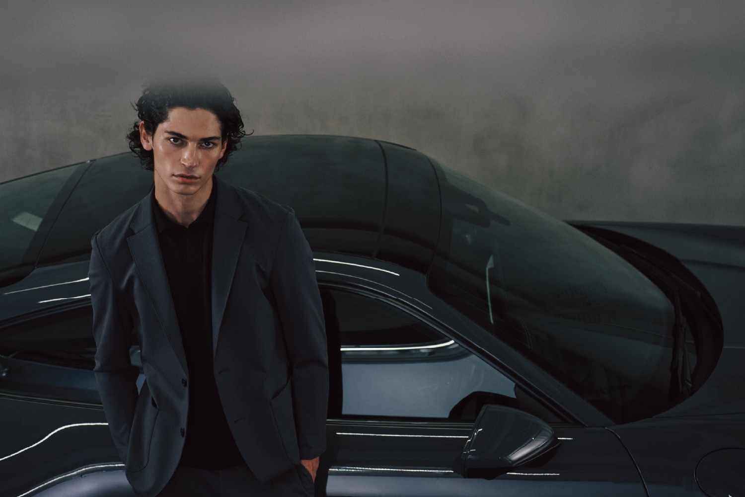 グレイのジャージーセットアップとブラックのポロシャツを着た男性が車の前に佇むビジュアル