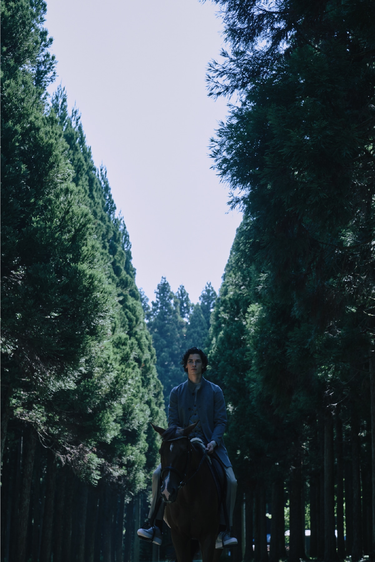 ライトグレーのウールスタンドコートとダウンジレとライトベージュのパンツを着用した男性が静謐な森の中で乗馬するビジュアル