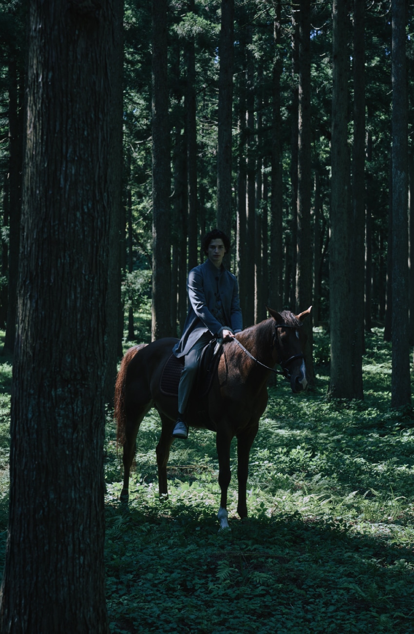 ライトグレーのウールスタンドコートとダウンジレとライトベージュのパンツを着用した男性が静謐な森の中で乗馬するビジュアル