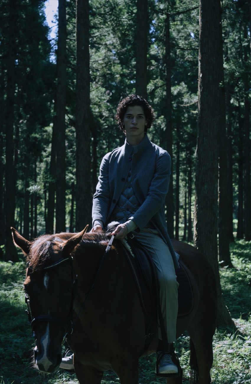 ライトグレーのウールスタンドコートとダウンジレとライトベージュのパンツを着用した男性が静謐な森の中で乗馬するビジュアル