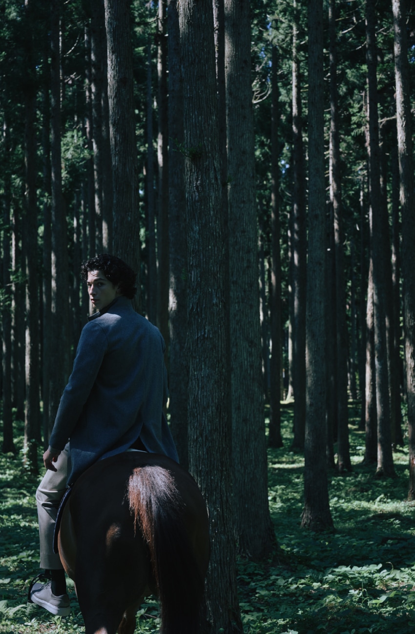 ライトグレーのウールスタンドコートとダウンジレとライトベージュのパンツを着用した男性が静謐な森の中で乗馬するビジュアル